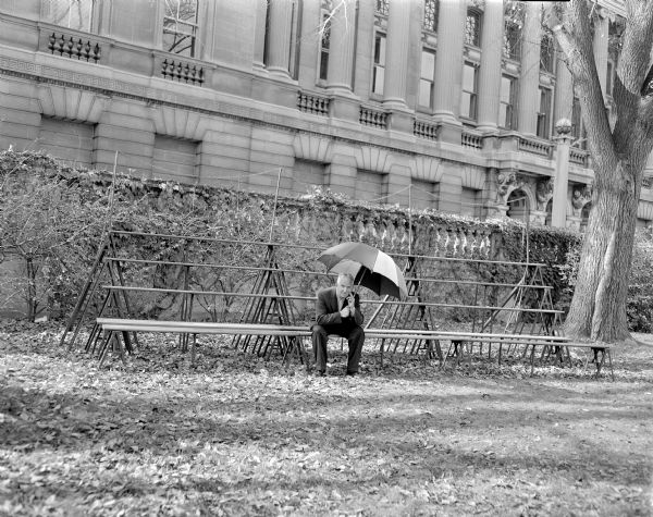 Sid sitting outdoors on bleachers on the Library Mall in front of the entrance to the Wisconsin Historical Society. He is holding an open umbrella above his head. 