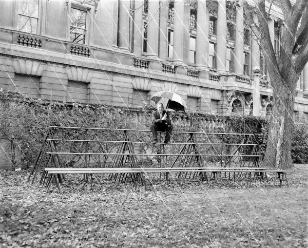 Sid sitting outdoors on the top row of a set of bleachers on the Library Mall in front of the entrance to the Wisconsin Historical Society. He is holding an open umbrella above his head. Streaks have been added to the negative to simulate rain.