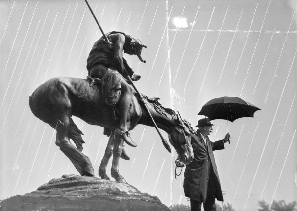 Sid posing with a cigar in his mouth while holding an umbrella above his head. He is standing next to the James Fraser bronze statue of a horse with an exhausted rider named <i>The End of the Trail</i>, while pretending to hold the bridle of the horse. Streaks have been added to the negative to simulate rain.