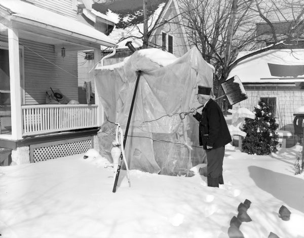 Sid standing in his snowy backyard looking at his wooden trolley, Toonerville, which has been covered with plastic for the winter. Some of Sid's sculptures are in the snow on the right. He is wearing eyeglasses, a dark coat, a railroad engineer style hat, and a corn cob pipe in his mouth. He is resting a snow shovel over his shoulder.