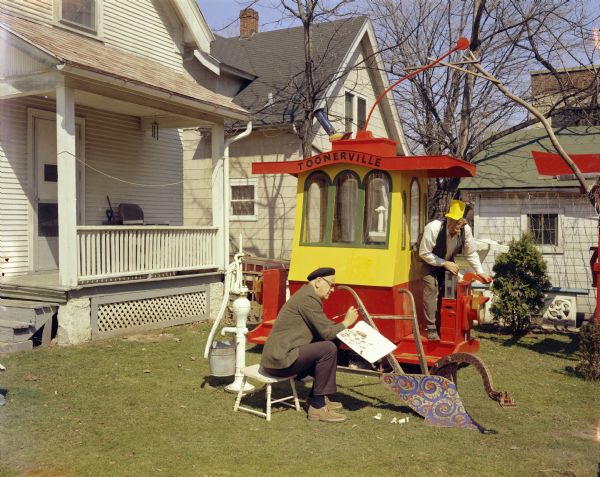View across the backyard of 237 Waubesa St. looking at Sid Boyum standing on the back of his wooden trolley, "Toonerville". Sid has a corncob pipe in his mouth and is wearing a yellow railroad engineer's-style hat. A man sitting in a chair in front of the trolley is using a paintbrush to decorate an old hand plow with paint. There is a hand-powered water pump with a bucket near the back porch.