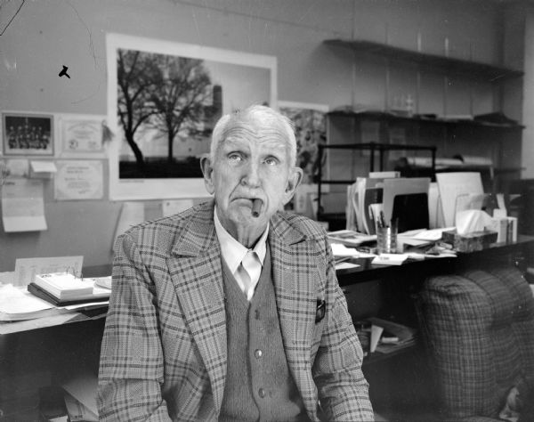 Waist-up portrait of Sid, looking upward, sitting in his office with his signature cigar in his mouth. He is wearing a sweater vest and a plaid jacket. The counter behind him is cluttered with papers.