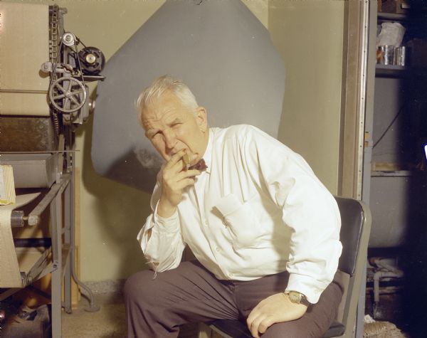 Portrait of Sid posing in the Gisholt darkroom while smoking a cigar. He is sitting in a chair and is wearing a ripped, button up white shirt with a bow tie. His fingernails are stained with what may be pyrogallol, a staining agent used in the development of film.