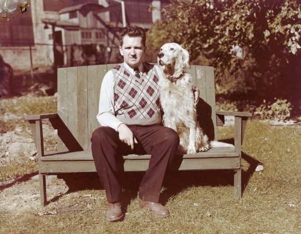 Portrait of Sid, wearing a sweater vest and a necktie, sitting outdoors in his backyard. He is posing on a bench with his arm around his dog, Duke, sitting next to him. In the background behind a fence is the Madison-Kipp Corporation.