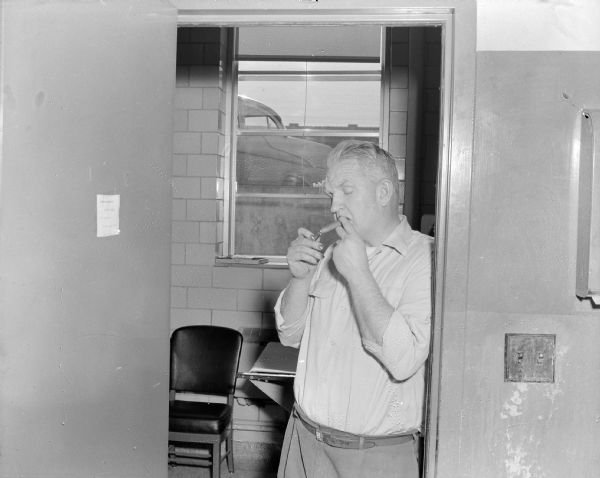 Sid standing and lighting up a cigar in the doorway of a room at work. Behind him is a table and chair, and outside of the window is a parked automobile. 