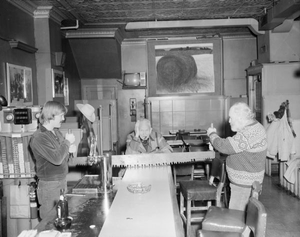 View down bar towards two men pretending to cut the bar in half with a large saw. One of the men stands behind the bar on the left, and the other man stands opposite him on the right. Sid is sitting on the other end of the bar watching with his head in his hand. 
