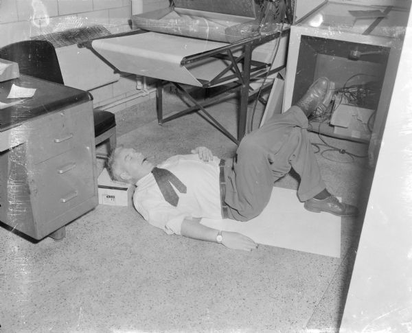 Sid sleeping on the floor near a desk in the darkroom at Gisholt with a cigar in his mouth. His legs are crossed and his head is resting on a cardboard box. 