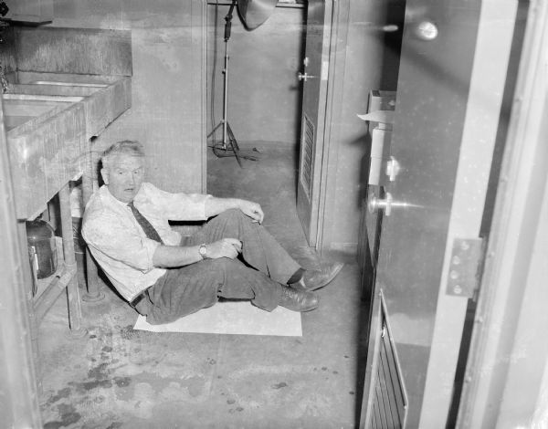 Sid is holding a cigar in his hand while sitting on the floor in the darkroom at the Gisholt Factory on a piece of paper near a sink, looking as if he just woke up from a nap.