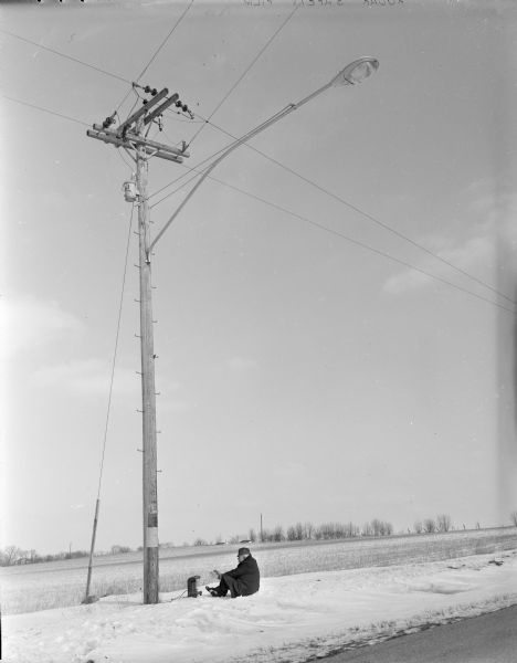 View from road of Sid sitting in the snow next to the base of a power pole while warming his hands by a small heater, which appears to be plugged into the power pole. In the background are fields and trees.