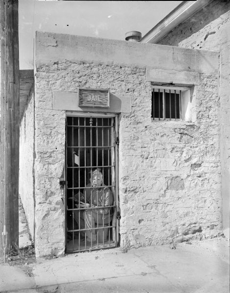 View from sidewalk of Sid sitting in jail with a cigar in his mouth while reading a Gideon Bible. Sid is looking out of the iron gate of a stone building, with a sign above the door reading: "JAIL." There is an iron barred window on the right.