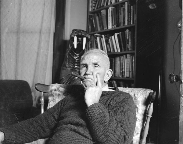 Sid sitting in a chair at home with his chin in his hand and a cigar in his mouth. A statue of a walrus and a bookcase full of books is in the background.