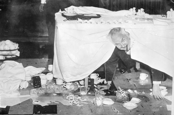 Sid caught underneath a table during a bridal dinner. Sid is looking out from under a tablecloth draped over his neck after trays of dishes crashed to the ground. He has a cigar in his mouth, while peering out over the floor covered in spilled cream and broken tea cups and saucers. In the background are the hands and legs of another man standing near the table.