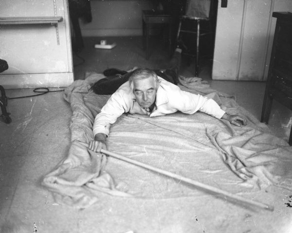An unknown man with his eyes closed is crawling on a cloth on the floor of a room. He is holding a stick in his right hand, and his left foot is caught on a stool in the background.