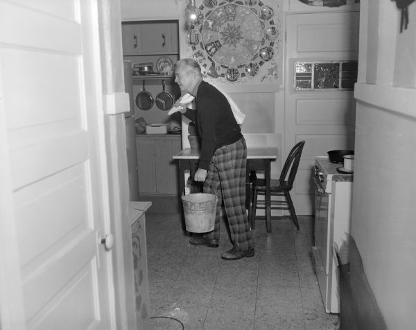 A man is posing in profile in Sid's kitchen, carrying a bucket in his left hand and holding a towel over his right shoulder. On the wall in the background is an artwork by Sid that has twelve plates, each with an astrological figure on it and a roman numeral from I-XII. They surround an interior design of more astrological designs.