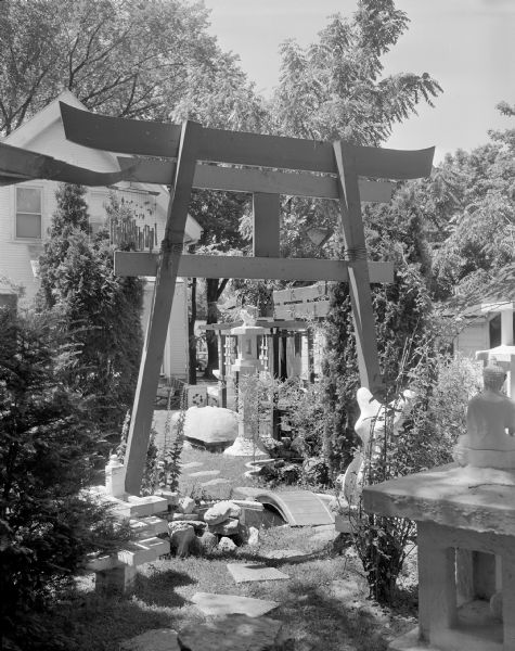 View of Sid's backyard looking towards the back of the house. There is a large arch in the center with wind chimes hanging on it. Shrubs, trees, plants and other sculptures are on either side of a path, which is paved with stones spaced in the lawn. There is a small arched wooden bridge over a pond directly underneath the arch. The edge of the roof of the pagoda is in the foreground on the left.