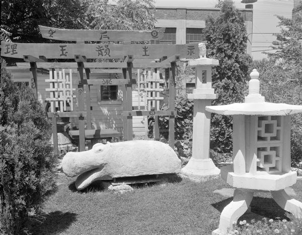 Black-and-white image of the torii gate several cast-concrete sculptures - two white lanterns (one being the "Fu Dog Lantern") and the "Hippopotamus" in Sid's backyard. The "Hippopotamus" measures 19" x 25" x 69" and the "Fu Dog Lantern" 85" x 19" in diameter. Behind the sculptures is a small wood building, perhaps a garage. The Madison-Kipp Corporation factory is in the far background.