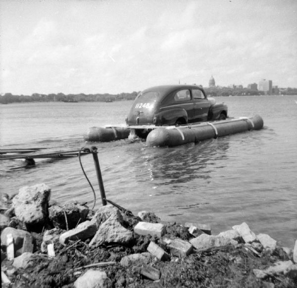 View from rocky shoreline of the launching of the "Monona-Mobile" (pontoon car) onto Lake Monona. The boat launch is on the left. "11248" is painted on the car's back. The Wisconsin State Capitol is on the far shoreline in the background. 

An article called "The 'Monona-Mobile'" was published in a 1955 issue of the <i>Gisholt Crib</i> explains: "First [John "Commodore" Heggestad] constructed a pontoon support for his car, then, on the rear tires, he chained wooden cleats which serve as paddles and propel the pontoon car at a respectable speed — as high as 10 knots per hour. Steering is accomplished by a rudder suspended from the rear axle. Gisholt lensman, Sid Boyum, who recently took an action photo of the skipper and his craft from the Lake Monona shoreline, reports that the 'cruiser' appears to be completely seaworthy."