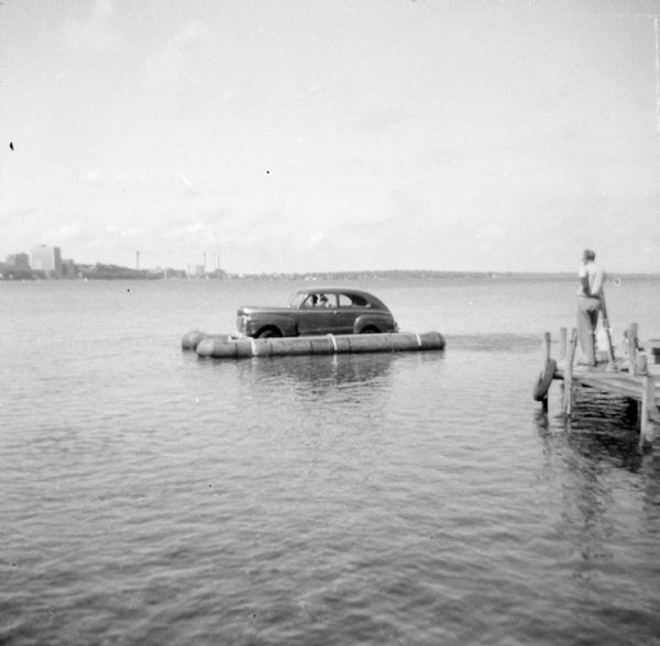 View across water towards the "Monona-Mobile" (pontoon car) on Lake Monona, close to a pier on the left where a man is standing and watching. Buildings on the isthmus are in the background.

An article called "The 'Monona-Mobile'" published in a 1955 issue of the <i>Gisholt Crib</i> explains: "First [John "Commodore" Heggestad] constructed a pontoon support for his car, then, on the rear tires, he chained wooden cleats which serve as paddles and propel the pontoon car at a respectable speed--as high as 10 knots per hour. Steering is accomplished by a rudder suspended from the rear axle. Gisholt lensman, Sid Boyum, who recently took an action photo of the skipper and his craft from the Lake Monona shoreline, reports that the 'cruiser' appears to be completely seaworthy."