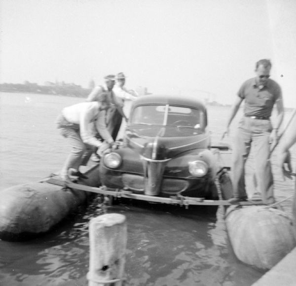 A group of men are standing on the pontoons of the "Monona-Mobile" (pontoon car) which is near a pier on the left. The Wisconsin State Capitol is in the far background. 

An article called "The 'Monona-Mobile'" published in a 1955 issue of the <i>Gisholt Crib</i> explains: "First [John "Commodore" Heggestad] constructed a pontoon support for his car, then, on the rear tires, he chained wooden cleats which serve as paddles and propel the pontoon car at a respectable speed--as high as 10 knots per hour. Steering is accomplished by a rudder suspended from the rear axle. Gisholt lensman, Sid Boyum, who recently took an action photo of the skipper and his craft from the Lake Monona shoreline, reports that the 'cruiser' appears to be completely seaworthy."