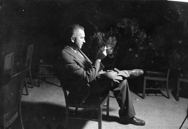 Portrait in profile of Sid Boyum sitting in the dark smoking a cigar while sitting in a chair.