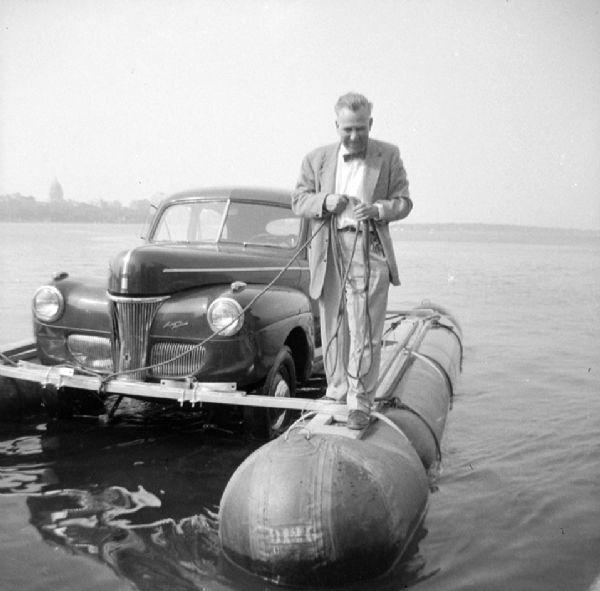 Unidentified man wearing a suit and a bow tie is standing on a pontoon of the "Monona-Mobile" (pontoon car) and holding the tow rope. Across the lake is the Wisconsin State Capitol. John "Commodore" Heggestad of Department 40 of Gisholt designed the method, the first man to cross Lake Monona by car on water.

An article called "The 'Monona-Mobile'" published in a 1955 issue of the <i>Gisholt Crib</i> explains: "First [John "Commodore" Heggestad] constructed a pontoon support for his car, then, on the rear tires, he chained wooden cleats which serve as paddles and propel the pontoon car at a respectable speed--as high as 10 knots per hour. Steering is accomplished by a rudder suspended from the rear axle. Gisholt lensman, Sid Boyum, who recently took an action photo of the skipper and his craft from the Lake Monona shoreline, reports that the 'cruiser' appears to be completely seaworthy."