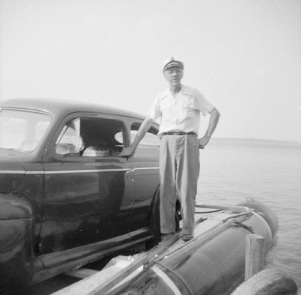 View from pier of John "Commodore" Heggestad of Department 40 of Gisholt proudly standing on the pontoon of the "Monona-Mobile" (pontoon boat) on which his car is mounted. He designed the method, the first man to cross Lake Monona by car on water.

An article called "The 'Monona-Mobile'" published in a 1955 issue of the <i>Gisholt Crib</i> explains: "First [John "Commodore" Heggestad] constructed a pontoon support for his car, then, on the rear tires, he chained wooden cleats which serve as paddles and propel the pontoon car at a respectable speed--as high as 10 knots per hour. Steering is accomplished by a rudder suspended from the rear axle. Gisholt lensman, Sid Boyum, who recently took an action photo of the skipper and his craft from the Lake Monona shoreline, reports that the 'cruiser' appears to be completely seaworthy."
