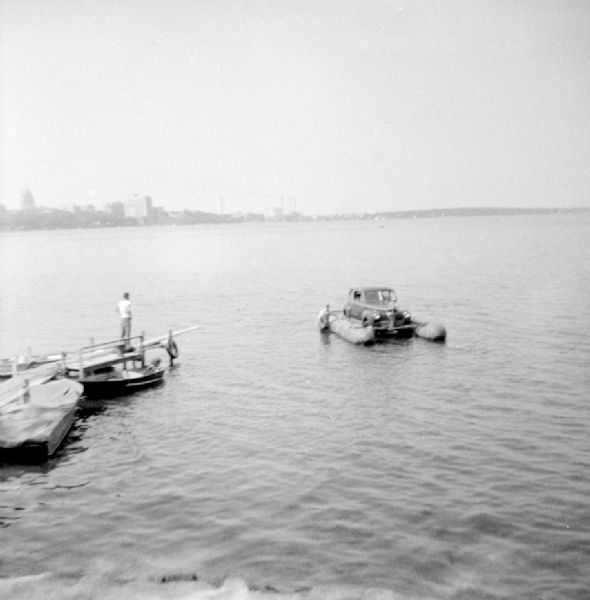 Elevated view from shoreline of the "Monona-Mobile" (pontoon boat) which is powered by an automobile. There is a man standing on a pier on the left watching another man who is standing in the water next to one of the pontoons. John "Commodore" Heggestad of Department 40 of Gisholt designed the method, the first man to cross Lake Monona by car on water. The Wisconsin State Capitol is in the far background.

An article called "The 'Monona-Mobile'" published in a 1955 issue of the <i>Gisholt Crib</i> explains: "First [John "Commodore" Heggestad] constructed a pontoon support for his car, then, on the rear tires, he chained wooden cleats which serve as paddles and propel the pontoon car at a respectable speed--as high as 10 knots per hour. Steering is accomplished by a rudder suspended from the rear axle. Gisholt lensman, Sid Boyum, who recently took an action photo of the skipper and his craft from the Lake Monona shoreline, reports that the 'cruiser' appears to be completely seaworthy."

