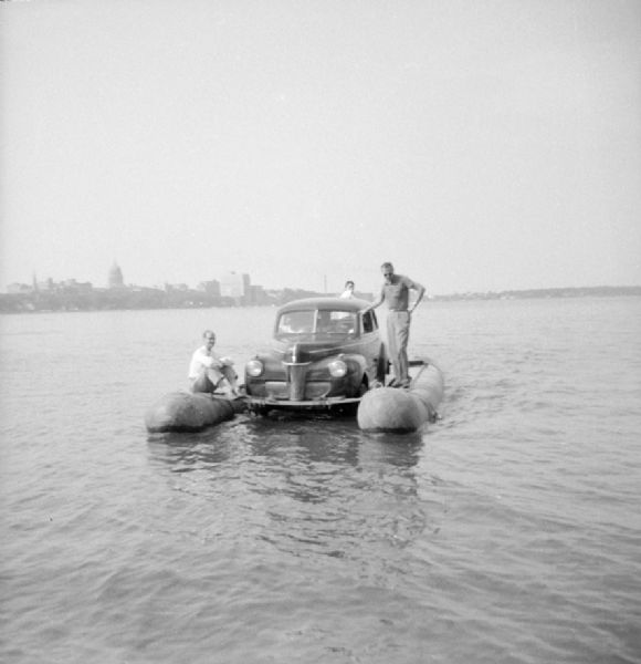 View across water of three men relaxing on the pontoons of the "Monona-Mobile." Two men are sitting in the front seat of the car. John "Commodore" Heggestad of Department 40 of Gisholt designed the method, the first man to cross Lake Monona by car on water. The Madison skyline, including the Wisconsin State Capitol, is in the background.

An article called "The 'Monona-Mobile'" published in a 1955 issue of the <i>Gisholt Crib</i> explains: "First [John "Commodore" Heggestad] constructed a pontoon support for his car, then, on the rear tires, he chained wooden cleats which serve as paddles and propel the pontoon car at a respectable speed--as high as 10 knots per hour. Steering is accomplished by a rudder suspended from the rear axle. Gisholt lensman, Sid Boyum, who recently took an action photo of the skipper and his craft from the Lake Monona shoreline, reports that the 'cruiser' appears to be completely seaworthy."