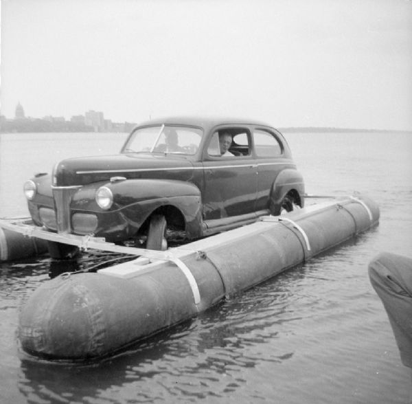 View from pier towards two men sitting in the front seat of the "Monona-Mobile," a pontoon boat powered by an automobile. The knee of a man is in the right foreground. John "Commodore" Heggestad of Department 40 of Gisholt designed the method, the first man to cross Lake Monona by car on water. The Madison skyline, including the Wisconsin State Capitol, is in the background.

An article called "The 'Monona-Mobile'" published in a 1955 issue of the <i>Gisholt Crib</i> explains: "First [John "Commodore" Heggestad] constructed a pontoon support for his car, then, on the rear tires, he chained wooden cleats which serve as paddles and propel the pontoon car at a respectable speed--as high as 10 knots per hour. Steering is accomplished by a rudder suspended from the rear axle. Gisholt lensman, Sid Boyum, who recently took an action photo of the skipper and his craft from the Lake Monona shoreline, reports that the 'cruiser' appears to be completely seaworthy."