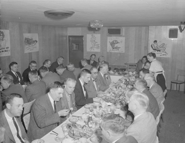Slightly elevated view of the "Ice Chippers," an ice fishing club of Gisholt that are at a dinner in a restaurant. The men are wearing suits and ties, and are seated at long tables. On the walls are hand-drawn cartoons with captions.