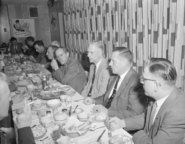 View down long table of members of the Ice Chippers of Gisholt having dinner, They are sitting in a restaurant, where comical hand-drawn drawings created by Sid hang on the walls. The man in the left foreground is holding a cigarette in his right hand, which is missing two fingers. 