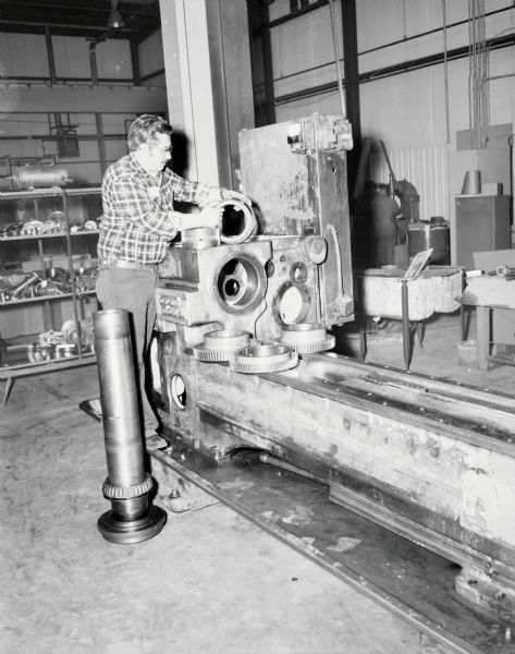 A man is standing and holding a part on top of a large piece of machinery inside the Gisholt factory. Behind him machinery parts are stored on shelving.