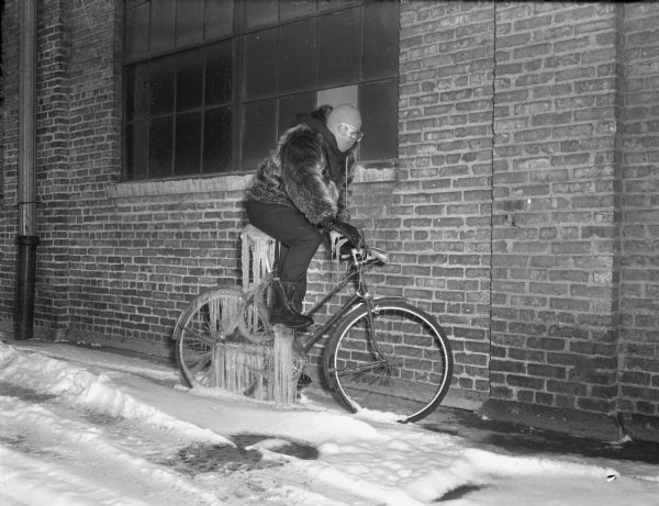 A man, wearing a fur coat, galoshes, a hat, and goggles is pretending to ride a bicycle that is covered by icicles. The bicycle is parked outdoors along the side of a brick building. Snow is on the ground. 
