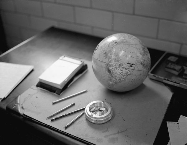 View of objects on a Gisholt desk: blotter, notepad, pencils, earth globe, and cigarette ends in an ashtray. 