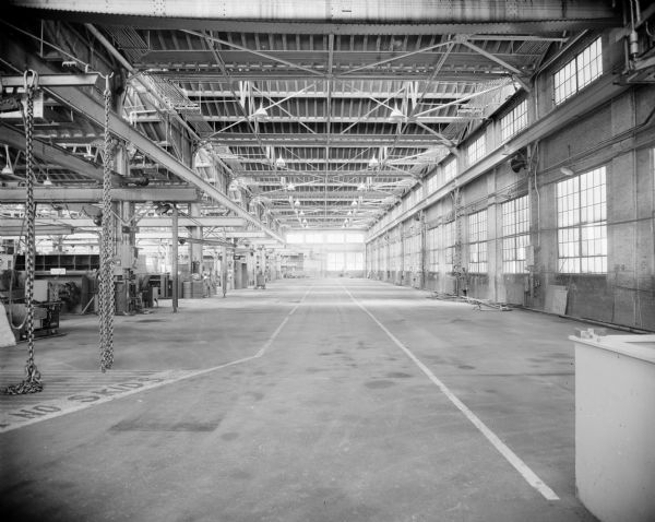 Interior view of the Gisholt factory, with exposed steel beams and large windows along the brick walls. On the left chains hang from hooks attached to beams, and a sign painted on the concrete floor reads: "No Skids."