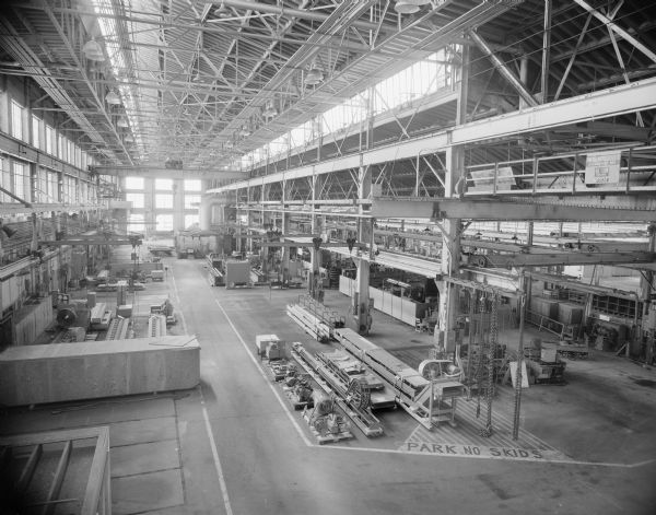 Elevated view looking down at the interior of the Gisholt factory. There is a large crane in the center of the room, and large windows line the walls. Machinery is packed on crates is sitting on the factory floor.