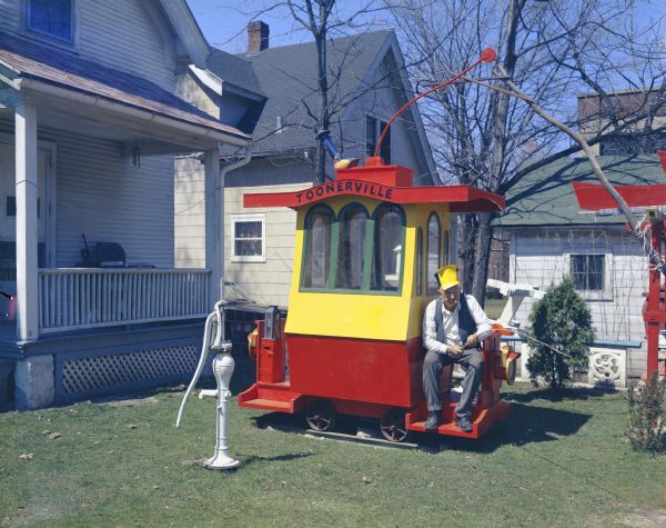 View of Sid sitting in his backyard on the back of his trolley car, "Toonerville," holding a stick in his hand and pretending to fish. Sid is wearing a large hat with a yellow bill, and has a cigar in his mouth. A hand water pump is in the yard on the left near the back porch. In the background is the old Communion rail from St. Bernard's Catholic Church and the house and garage next door.
