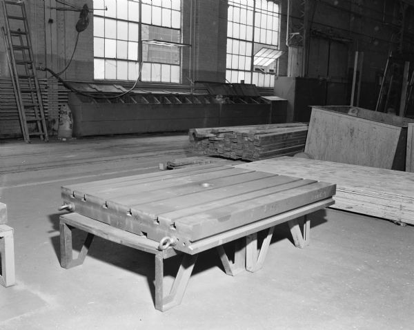 Interior view in the Gisholt factory of a large machinery part on a low stand. There are stacks of wood and lumber on the right, and windows are open in the background.