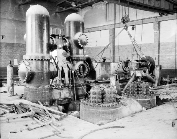 A man is welding on a large machine inside the Gisholt factory. On the right a crane is holding another section of the machine.