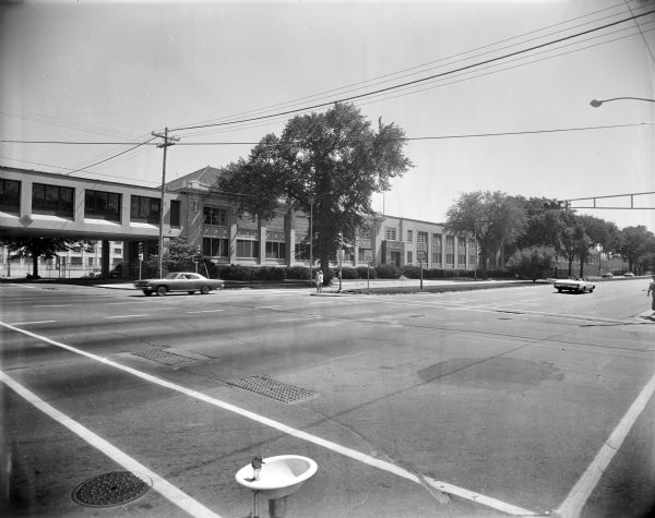 View from corner of East Washington Avenue at Baldwin Street towards the Gisholt factory. In the foreground is a water fountain (bubbler) on the sidewalk. There is an enclosed skybridge (called an "overhead" at the time of the industrial expansion approval in 1966) that connects the factory buildings across Baldwin Street.