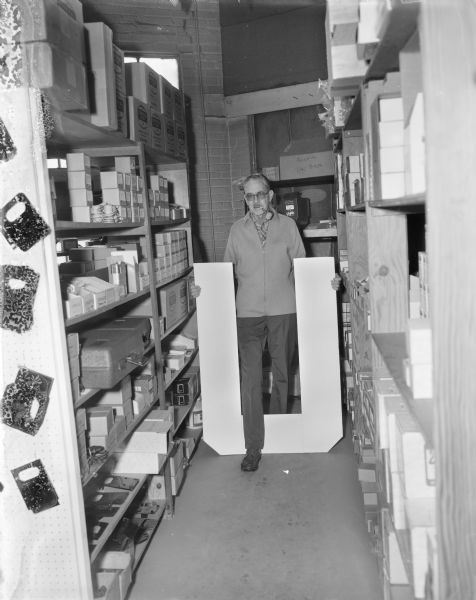 Portrait of a man with a pipe in his mouth posing while standing between storage shelves holding a large piece of cardboard or paper in the shape of a "U." He has his right leg stepping forward through the center of the "U."