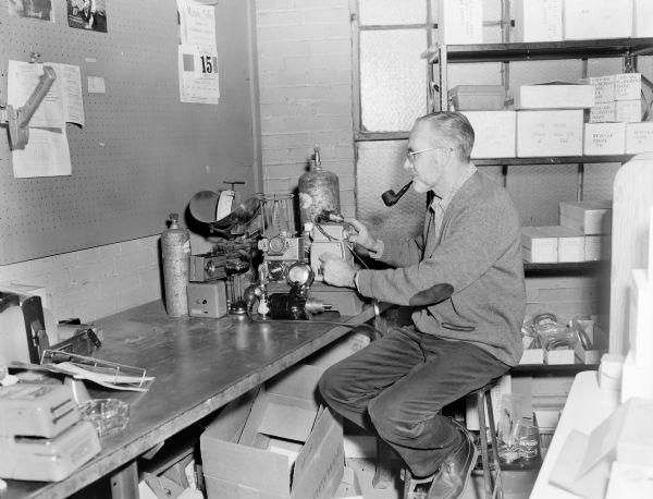 NEW:
View of a man sitting on a stool at a work table with a piece of machinery, probably at the Gisholt factory. He is wearing eyeglasses and is smoking a pipe. On the floor under the table are empty cardboard boxes, and on the right are shelves stacked with cardboard boxes, some marked: "Air Tube Combination."