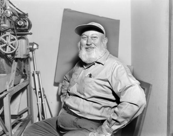 Waist-up portrait of an unidentified man sitting and smiling while posing next to a piece of machinery on a metal stand at the Gisholt factory. There is a piece of paper attached to the wall behind him, perhaps for a backdrop. There is a metal tripod leaning against the wall behind the machinery on the left.