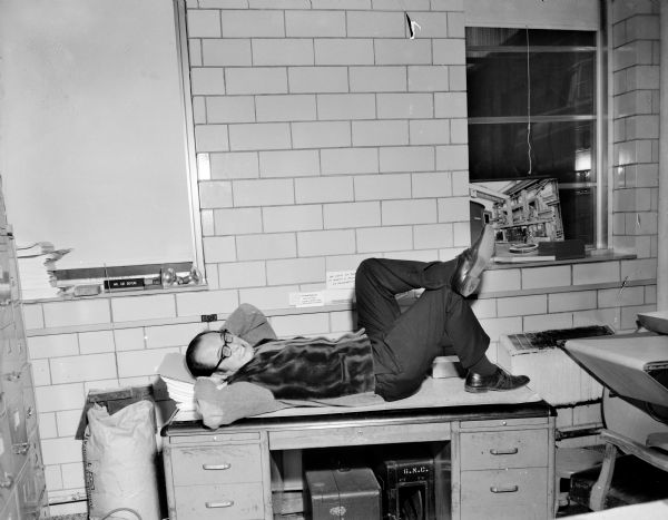Portrait of a man reclining with his head on his arms on top of a desk in the darkroom area at the Gisholt offices. Behind him on a windowsill is a desk nameplate that reads: "Mr. Sid Boyum." There are few sayings posted on the brick wall behind the desk, with one reading: "Accountability Profit or Loss?," "What Is Money — Money Talks Now What Were You Saying?," and "An ounce of today is worth a pound of tomorrow." On the windowsill on the right is propped a photograph of a factory, perhaps the Gisholt factory.