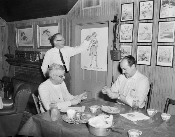 Two men from Ducks Unlimited are playing cards at a table, while another man is looking at a poster created by Sid titled: "Secrets of a Sweater Girl." The man is lifting a piece of paper on the poster, which exposes the woman's breasts which are tied in a knot. There are two framed images of ducks above the fireplace, and a set of nine framed images of ducks behind the table.