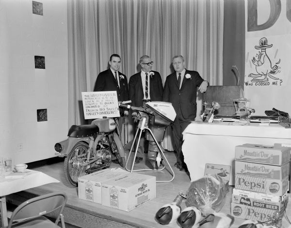 Three men are standing on a stage near a display of prize items to be given away during a Ducks Unlimited event. The Harley-Davidson motorcycle was donated by Decker H-D Sales and Harley-Davidson Mfg. Co. Other items on display are boxes of Pennzoil Outboard Motor Oil, a Johnson outboard motor, cases of Mountain Dew and Pepsi, and decoy ducks.