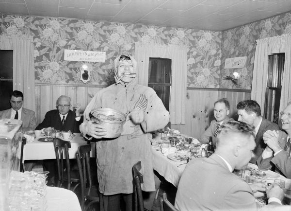 A man wearing rain gear is holding a pot of seafood with a fish in his mouth while standing in the center of tables during a Ducks Unlimited dinner. Men are eating in the background and on the right. A sign on the back wall reads: "Lampreys are just no dam good."
