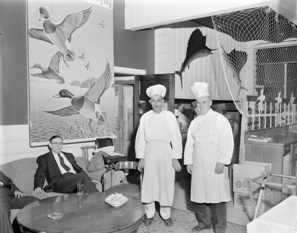 Two chefs for a Ducks Unlimited dinner standing and posing for a portrait. Men are sitting on a couch on the left under a large painting of ducks. People are working in the kitchen in the background. On the wall above the kitchen are two flying fish, and a large fishing net is suspended from the ceiling on the right.