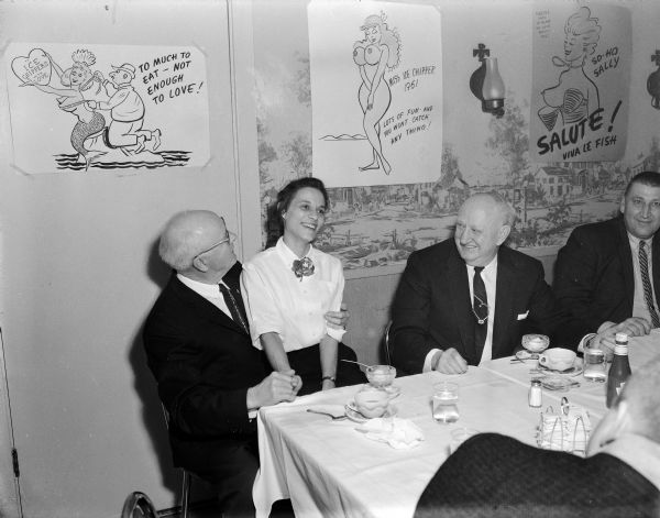 A woman sitting on the lap of a man at an Ice Chippers event. Several more men are sitting at the table. Posters are taped to the wall behind the group, and include "Miss Ice Chipper 1961" and "So-Ho Sally." 