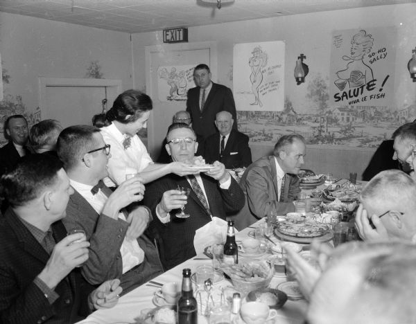 A woman is standing and serving dinner to men sitting at long tables at the Ice Chippers meeting. Posters are taped to the wall behind the group, and include "Miss Ice Chipper 1961" and "So-Ho Sally." 